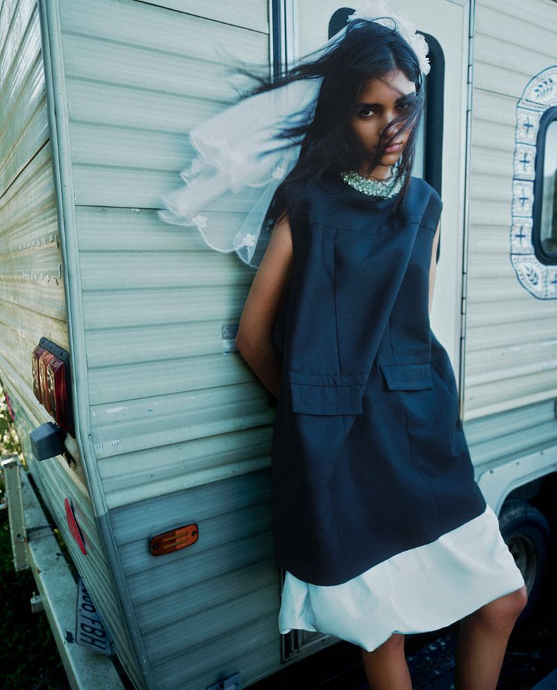 Model leaning against a pale caravan wearing a dark sleeveless tunic over a white skirt with a white veil in her hair