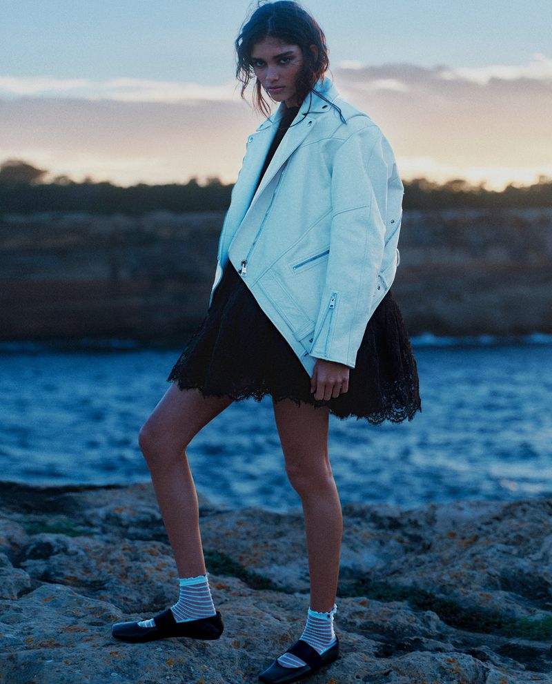 Model wearing a white leather jacket and black dress standing on rocky coast by the ocean at dusk