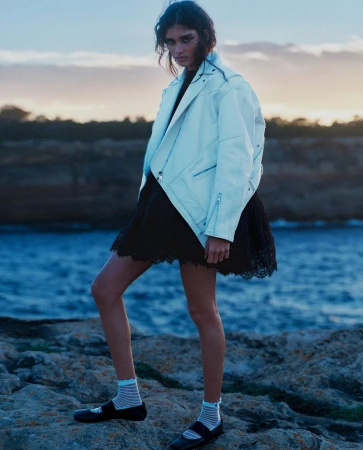 Model wearing a white leather jacket and black dress standing on rocky coast by the ocean at dusk