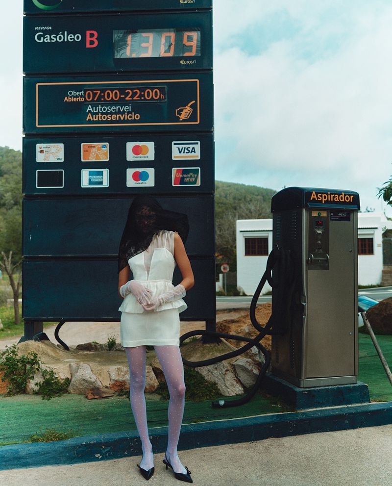 Model in a white minidress and sheer veil standing in front of a service station price sign and vacuum machine