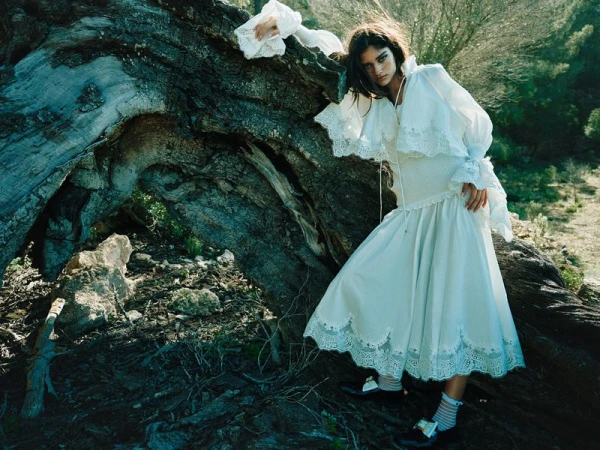 Model wearing a white ruffled dress leaning against a large hollow tree trunk in a forested landscape