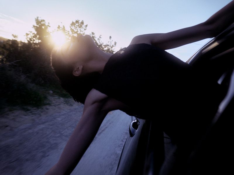 Model leaning out of car window with arms outstretched against a backlit sunset sky and roadside trees.