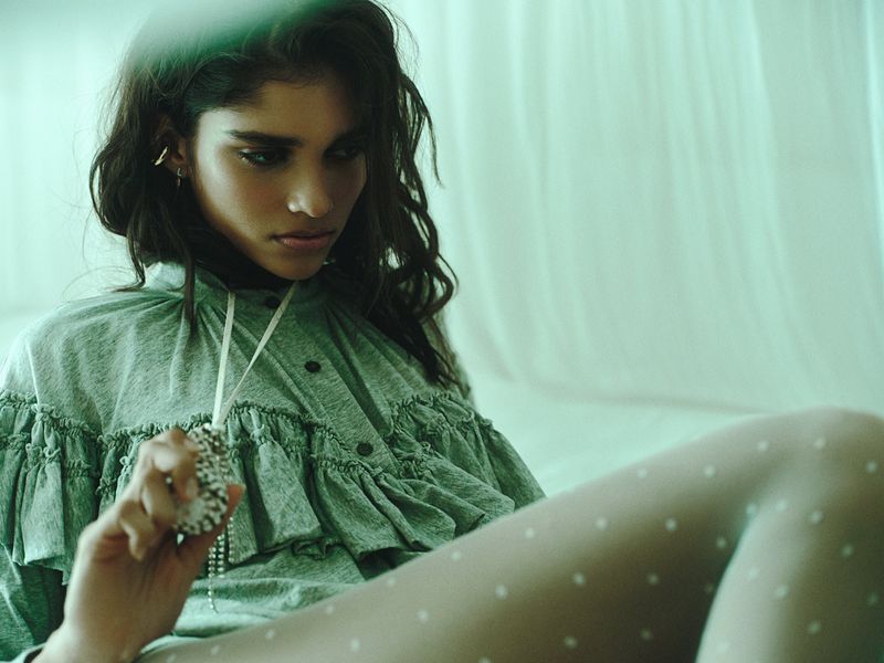 Model wearing a ruffled green blouse and dotted tights holding a decorative pendant against a draped backdrop