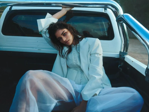 Model wearing a white structured blazer and sheer blue trousers sitting inside the back of a white pickup truck.