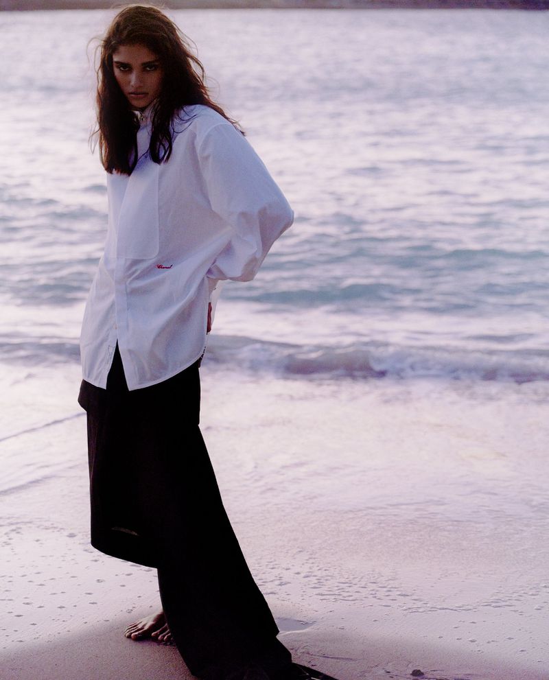 Model standing on a sandy beach wearing an oversized white button-down shirt and dark skirt by the ocean.