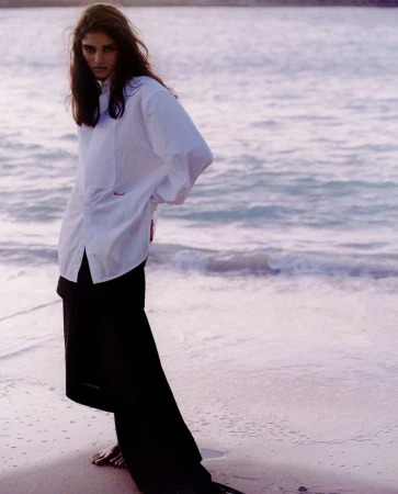 Model standing on a sandy beach wearing an oversized white button-down shirt and dark skirt by the ocean.