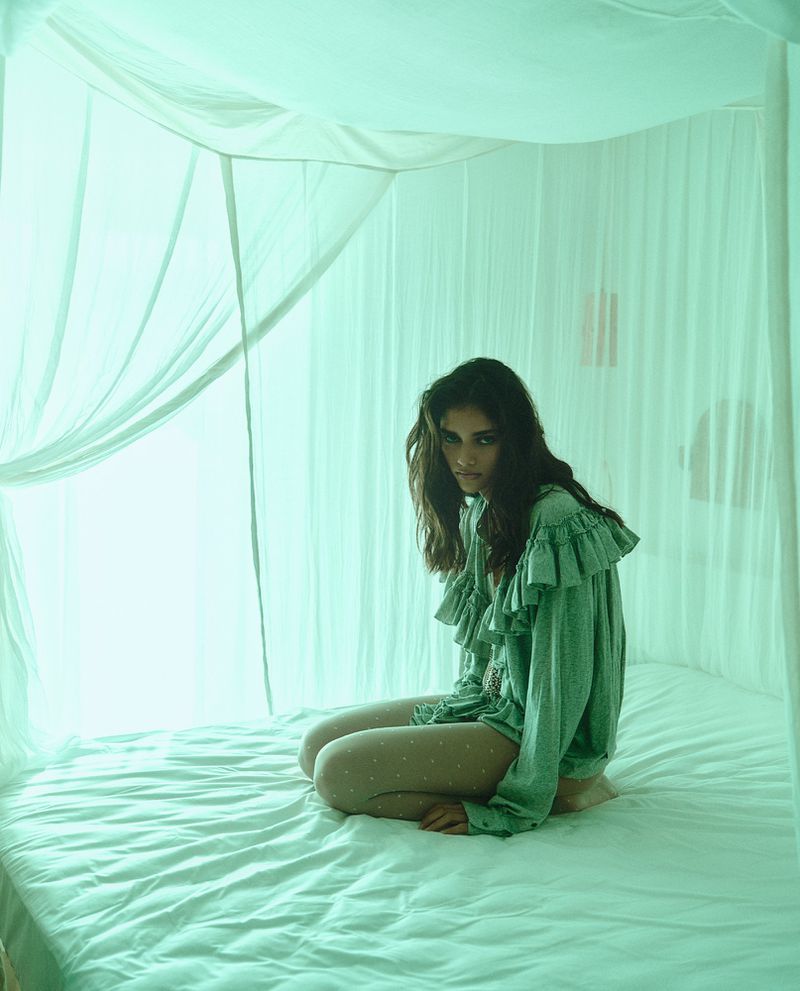 Model kneeling on a white canopied bed wearing a ruffled green blouse and sheer tights, enveloped in soft teal lighting.