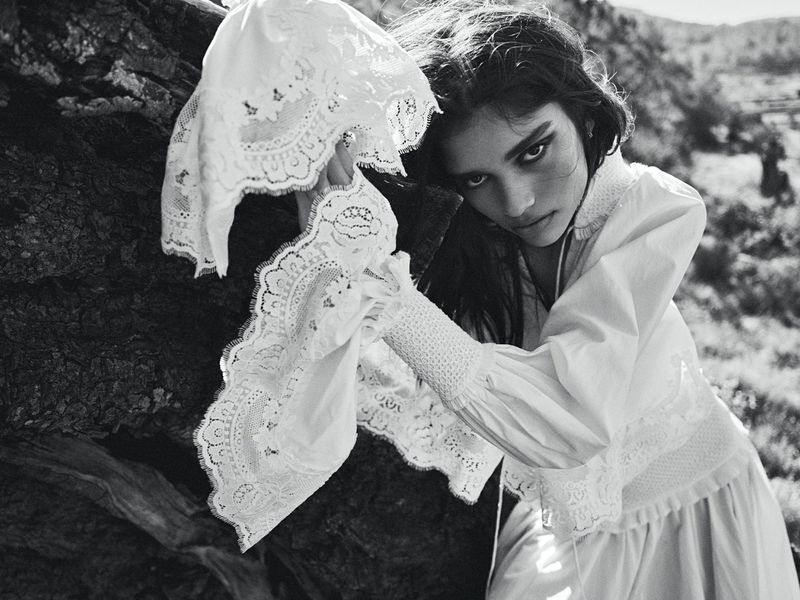 Black and white portrait of a woman in a white lace blouse posing against a dark tree trunk outdoors for Vogue Netherlands.