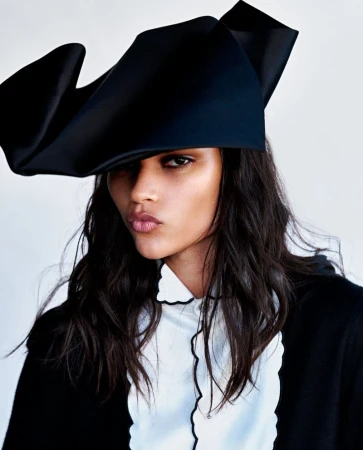 Model wearing a large black sculpted hat and white ruffled blouse against a plain light background.