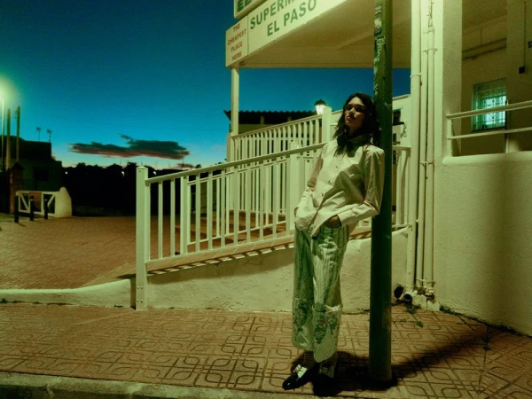 Model leaning against a green pole at dusk in front of a building with a white railing and tiled pavement.