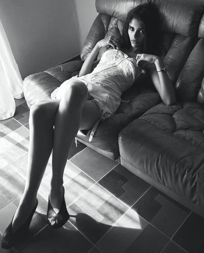 Model recline on a dark leather couch in a white lace dress with sunlight streaming across tiled floor.