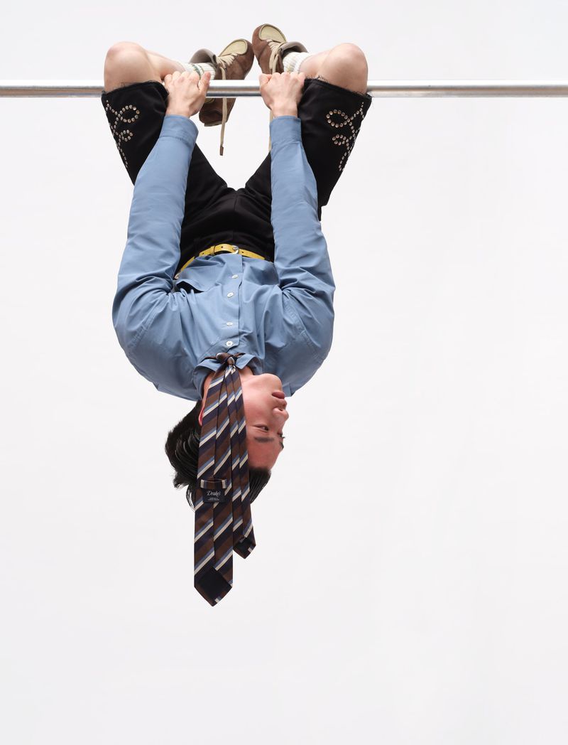 Model hanging upside down from a metal bar wearing a blue shirt and striped tie against a white backdrop
