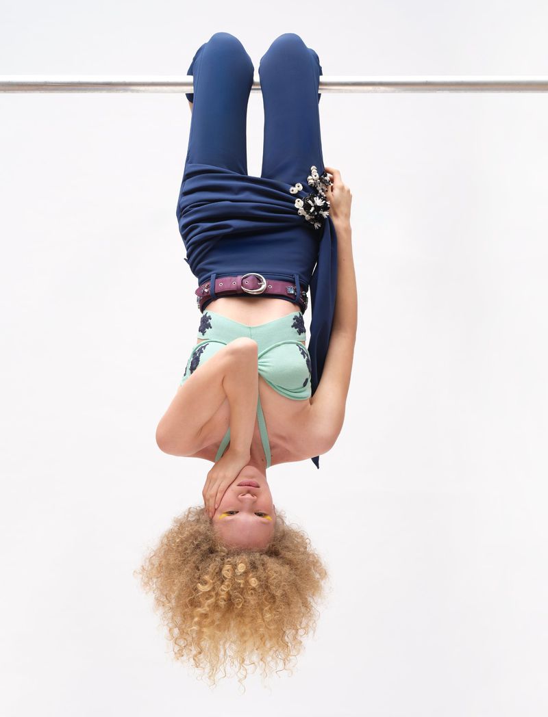 Upside-down woman with curly hair wearing a blue top, pants, and belt holds onto a metal bar against a bright white background.