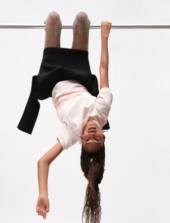Model hanging upside down from a metal bar wearing a pink t-shirt and black skirt against a white studio background