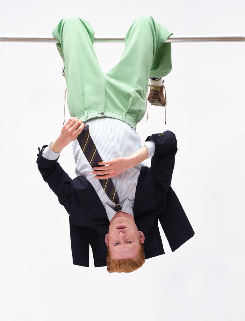 Man hanging upside down from a bar wearing a dark blazer, striped shirt, tie, and light green trousers against a white background.