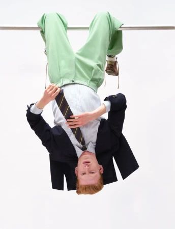 Man hanging upside down from a bar wearing a dark blazer, striped shirt, tie, and light green trousers against a white background.