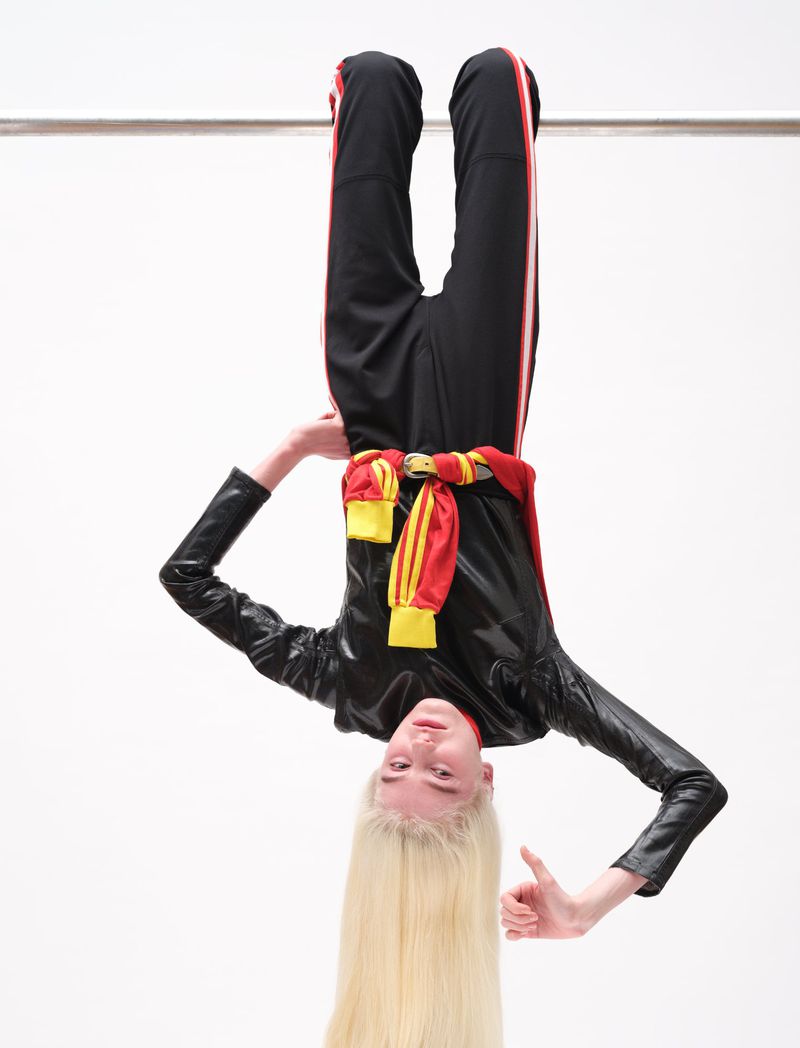 Model hanging upside down from a horizontal bar wearing a black track suit and red sweater tied around the waist against a white background