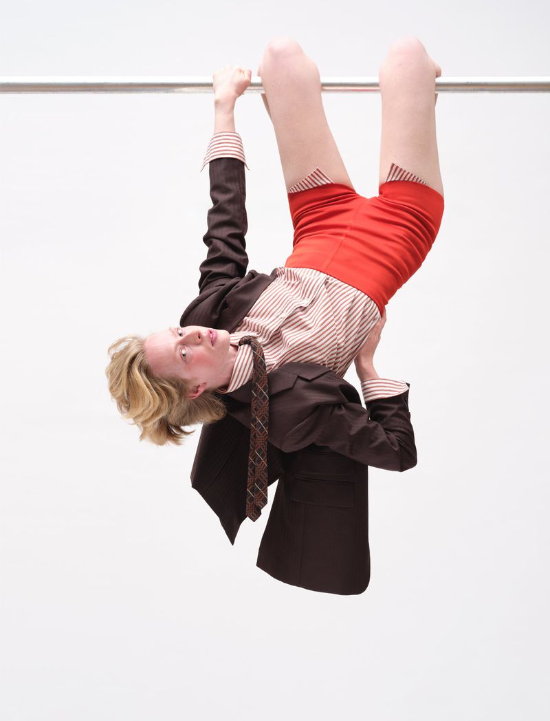 Model hanging upside down from a horizontal bar wearing a brown jacket, red shorts, and a patterned tie