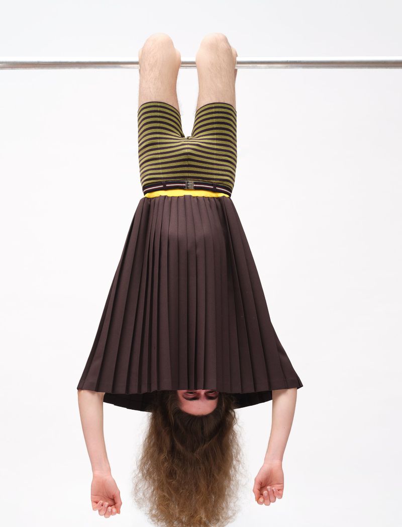 Model hanging upside down from a metal bar, wearing a brown pleated skirt and striped shorts against a plain white background.