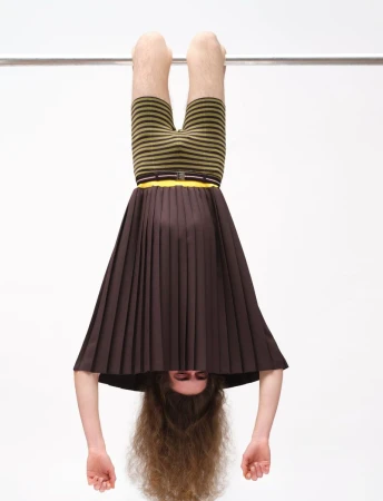 Model hanging upside down from a metal bar, wearing a brown pleated skirt and striped shorts against a plain white background.