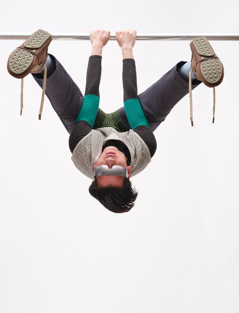 Model hanging upside down from a metal bar wearing sunglasses, layered knit vest, and brown lace-up shoes against a plain background
