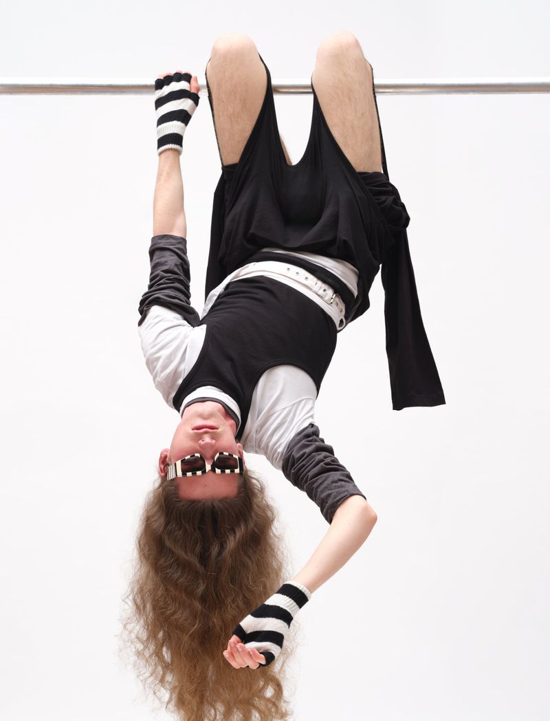 Model hanging upside down from a metal bar wearing black clothing and striped gloves against a white studio background.