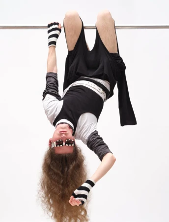 Model hanging upside down from a metal bar wearing black clothing and striped gloves against a white studio background.