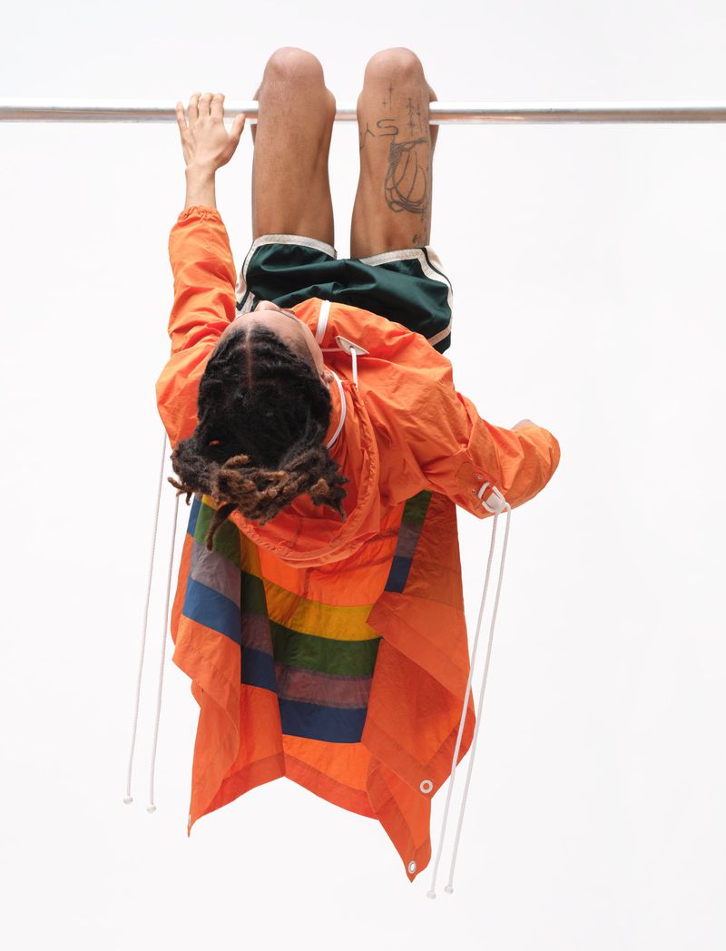 Person hanging upside down from a metal bar wearing an orange jacket and green shorts against a plain white background