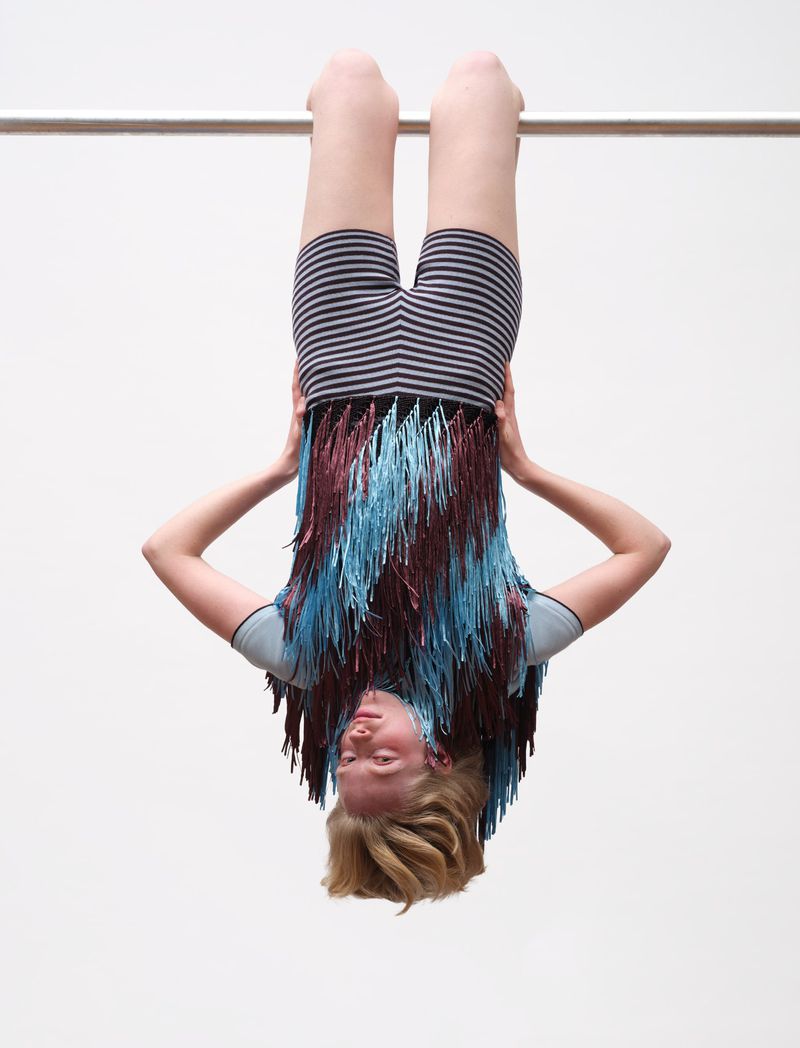 Model hanging upside down from a metal bar wearing striped shorts and a fringed top against a plain background