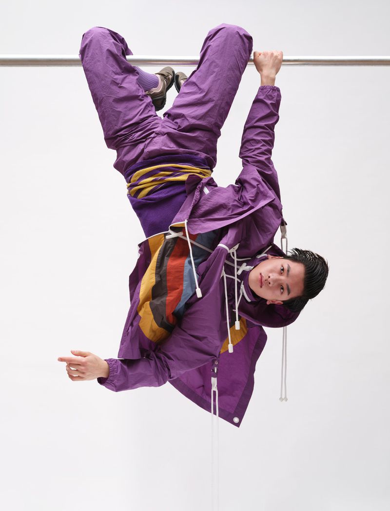 Model hanging upside down from a metal bar wearing a purple windbreaker and matching pants against a white background