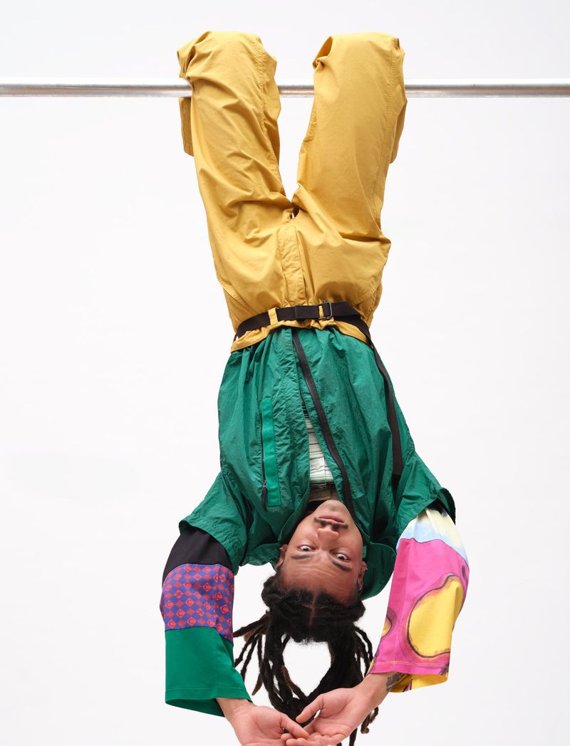 Model hanging upside down from a bar wearing a green jacket and yellow trousers against a white background