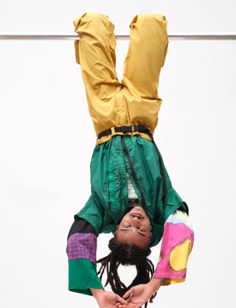 Model hanging upside down from a bar wearing a green jacket and yellow trousers against a white background