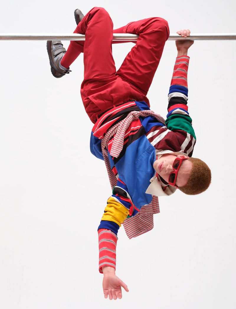 Model hanging upside down from a horizontal bar wearing colourful layered clothing against a plain white background.
