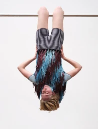 Model hanging upside down from a metal bar wearing striped shorts and a fringed top against a plain background