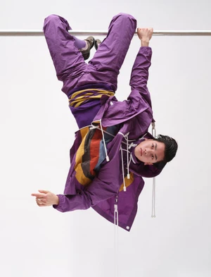 Model hanging upside down from a metal bar wearing a purple windbreaker and matching pants against a white background