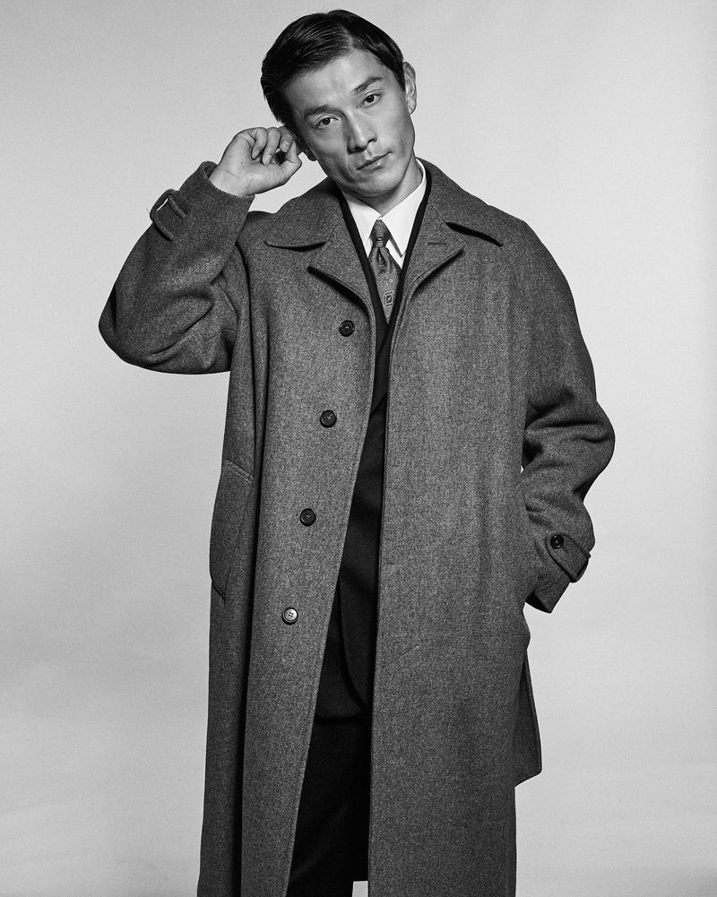 Monochrome portrait of a male model wearing a wool coat and tie with hand raised against a light studio backdrop.