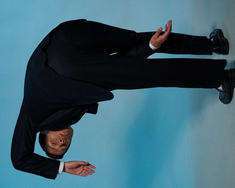 A man in a suit bends over backwards, his face and hands visible against a light blue background. The pose showcases flexibility.