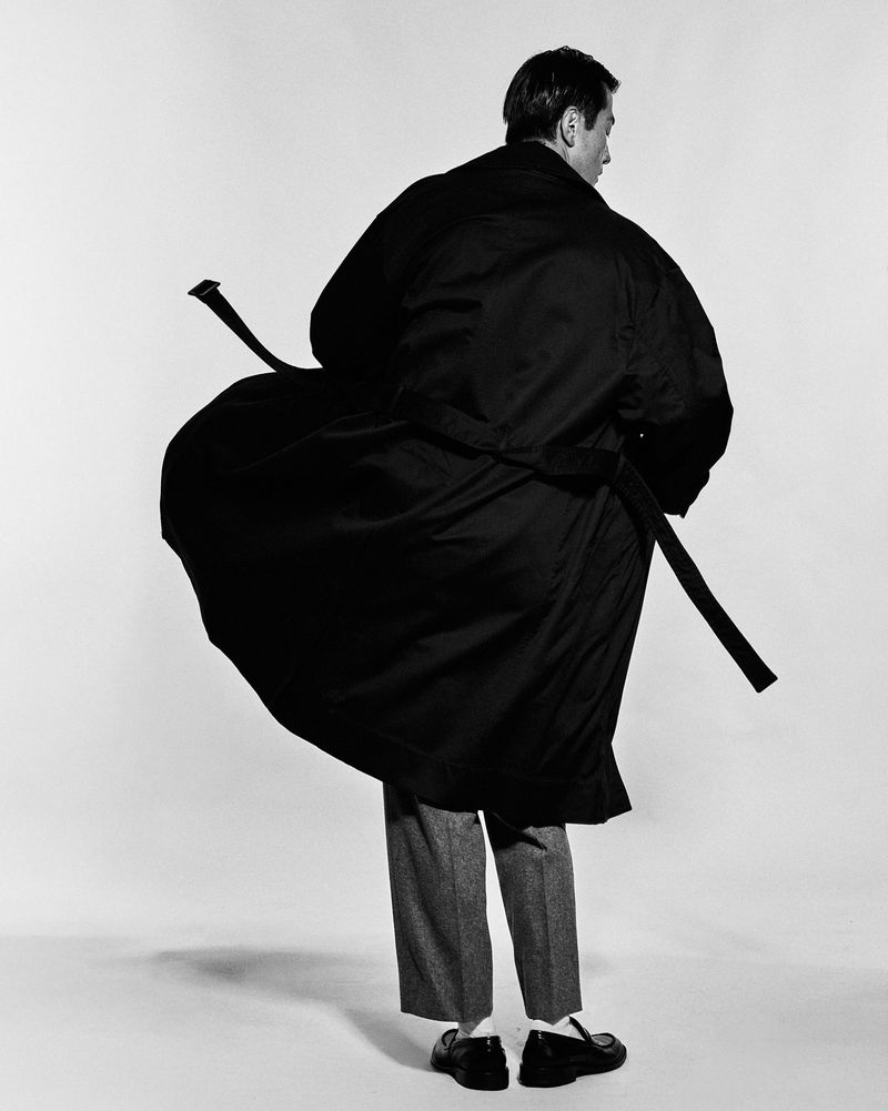 Man wearing a long dark belted coat and grey trousers viewed from behind against a white seamless background