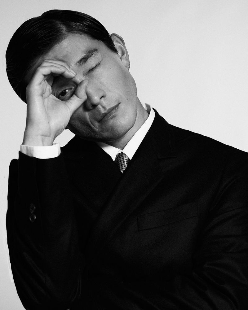 Man wearing a dark suit and tie with one hand raised to his face against a white studio background.