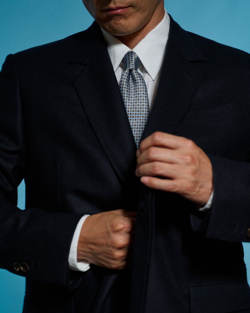 Close-up of a man wearing a dark tailored suit, white dress shirt, and patterned tie against a blue backdrop.