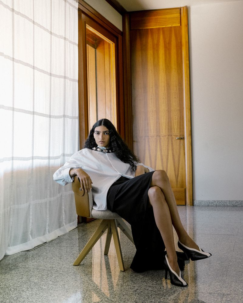 Model with long dark hair seated in a swiveling chair wearing a white shirt and black skirt against a wood panel wall