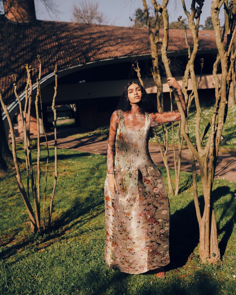 Model wearing patterned floral dress standing among bare trees on a grassy lawn in front of a modern architectural building