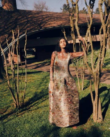 Model wearing patterned floral dress standing among bare trees on a grassy lawn in front of a modern architectural building