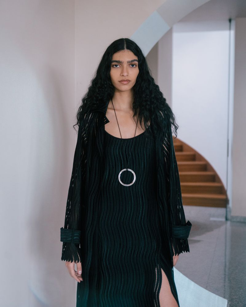Model Anita Pozzo wearing a dark ribbed dress and long black coat with a round pendant necklace indoors.