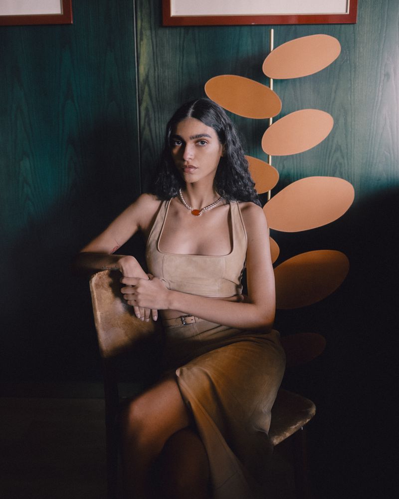 Model seated on a wooden chair wearing a beige top and skirt against dark green wood panelling with a decorative orange oval sculpture