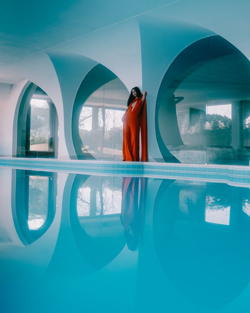 Model wearing a flowing orange gown standing by a swimming pool with dramatic arched architecture reflecting on the water.