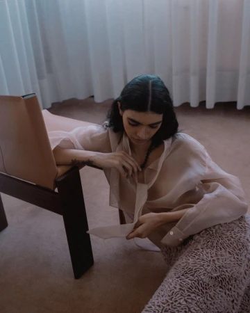 Model wearing sheer blouse and textured skirt seated on the floor leaning against a modern wooden chair.