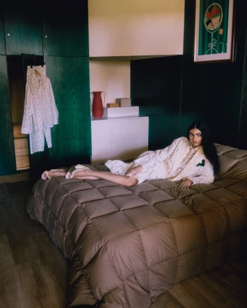 Model lying on a quilted brown bed inside a bedroom with dark green wood paneling, wearing cream-colored patterned apparel.