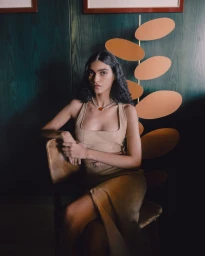 Model seated on a wooden chair wearing a beige top and skirt against dark green wood panelling with a decorative orange oval sculpture