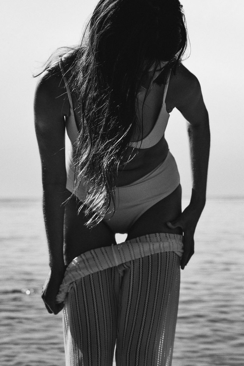 Black and white photograph of a woman pulling ribbed knit pants over a two-piece swimsuit while standing in the sea.
